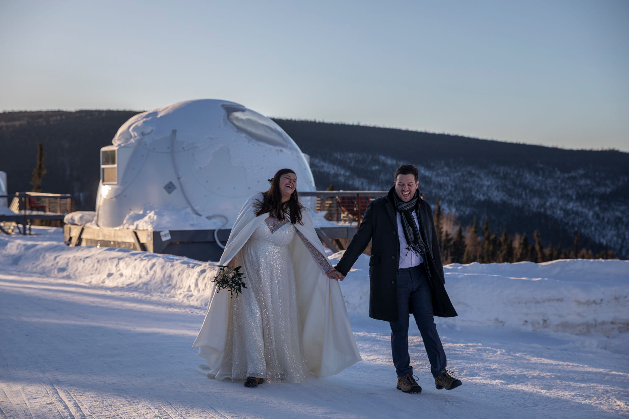 A bride and groom laugh walking hand in hand on a snowy path with a white igloo dome behind them.
