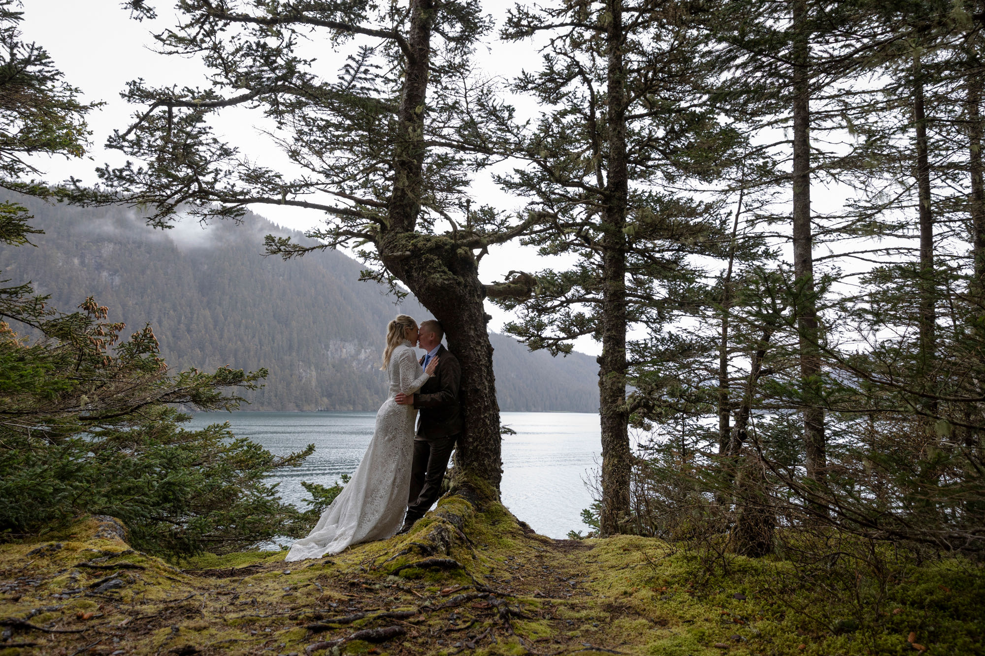 A groom leans against a tree in a mossy green forest in Alaska while his bride leans into him, giving him a kiss. 