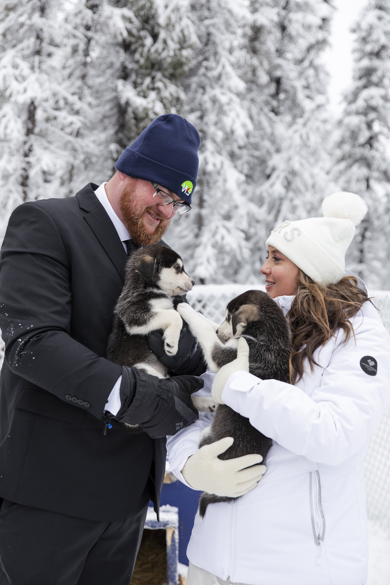A bride and groom wearing winter clothes, hats and gloves, hold a puppy in each other hands after they decided the best season to get married in Alaska was winter. 