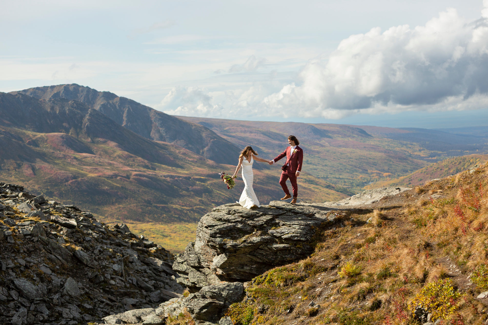 A bride and groom hold hands as they stand on top of a rock with beautiful views behind them.