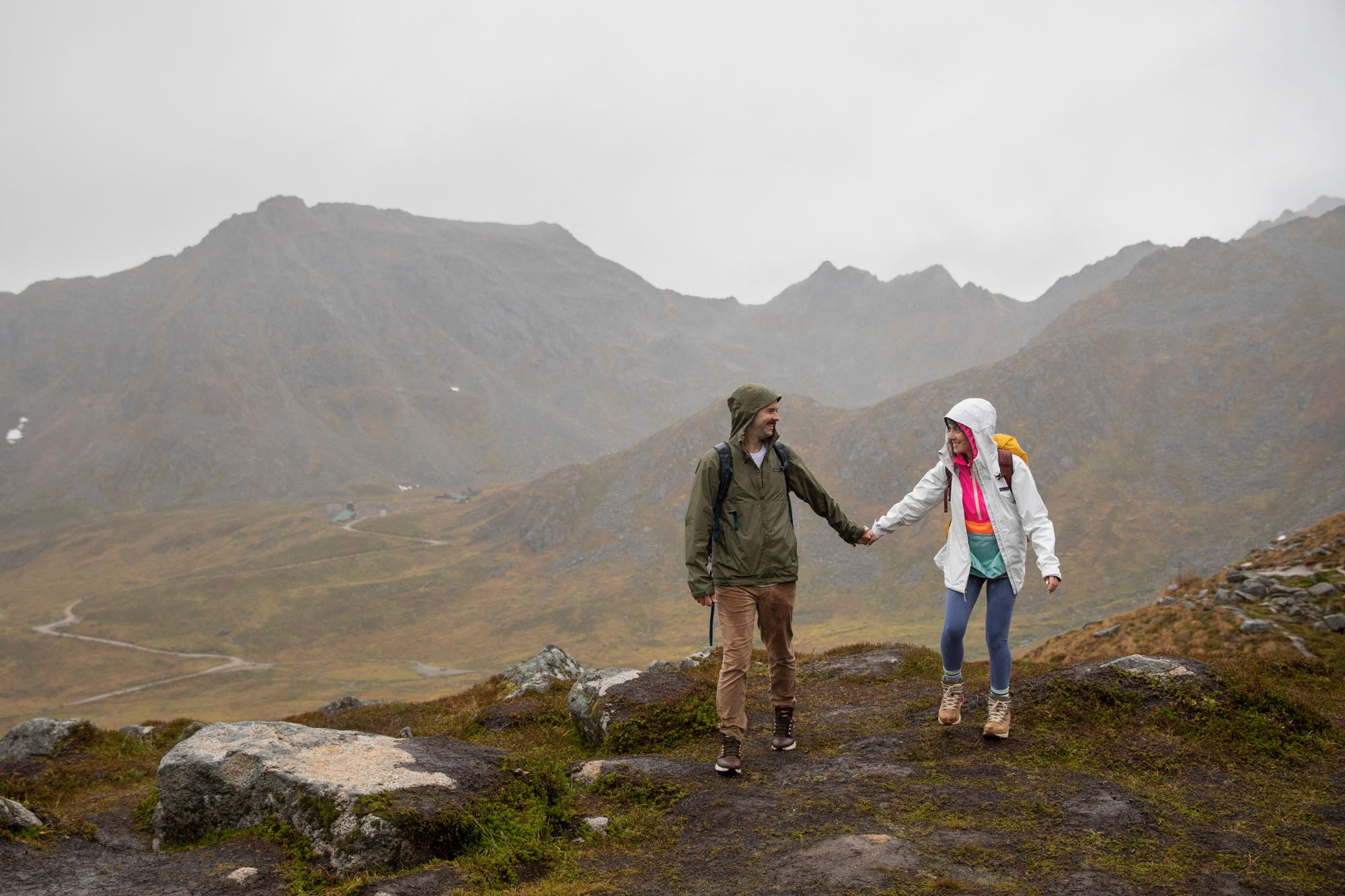 A couple is wearing rain jackets, holding hands on a hiking trail in Alaska. 