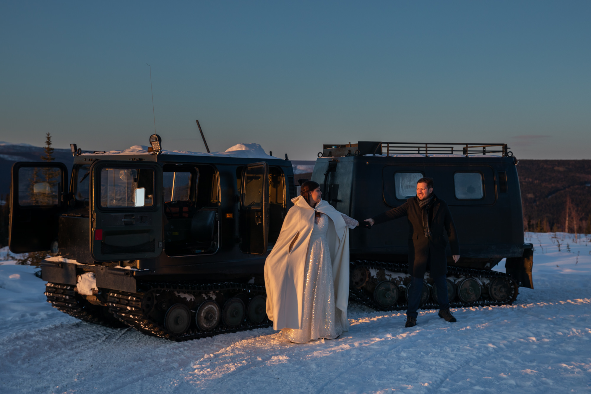 A bride wearing a white dress and white winter cape holds hands with her groom who is bundled up for winter on their Borealis Basecamp elopement day.