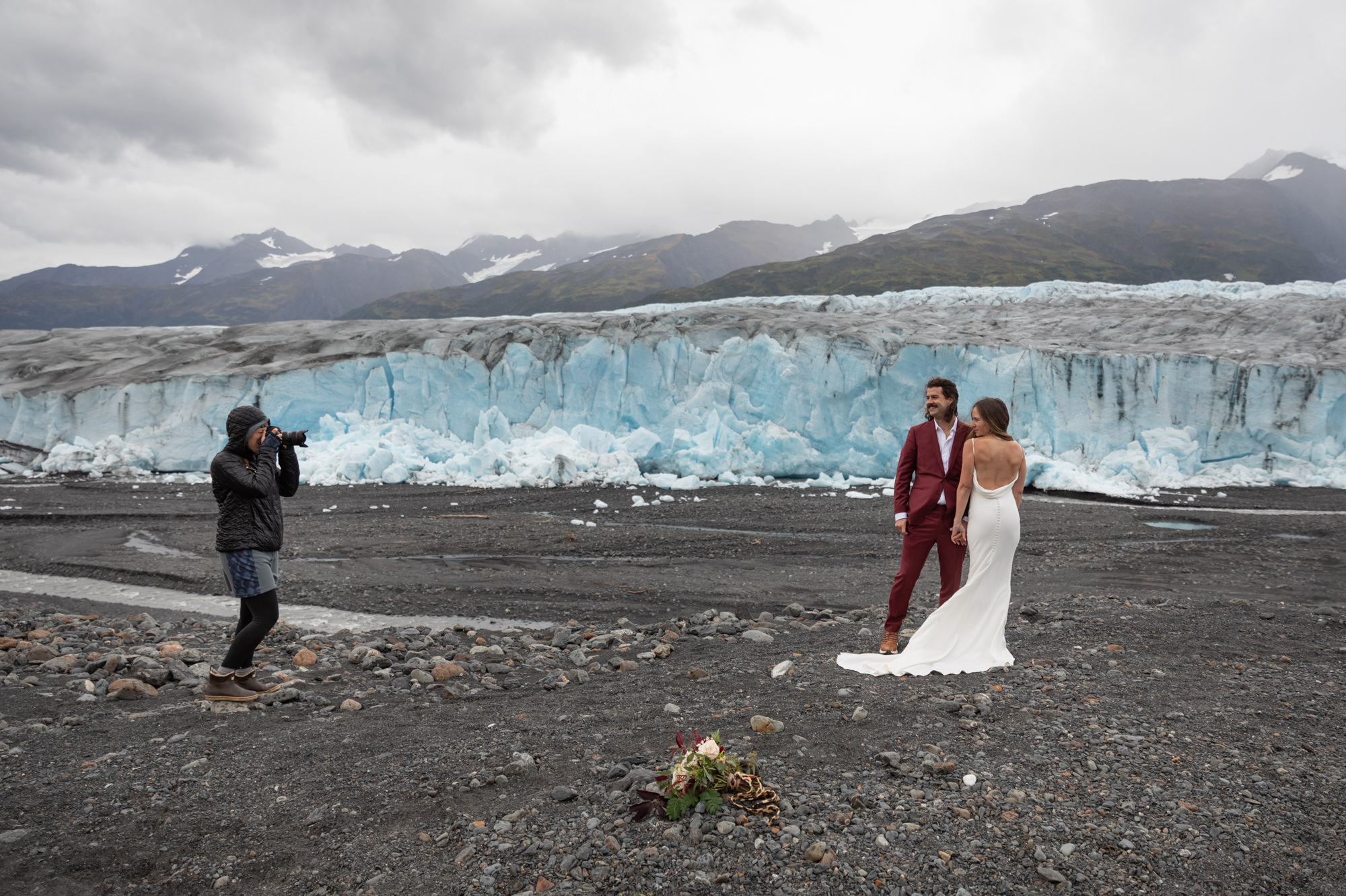 A couple dressed for their elopement poses in front of a stunning glacier, with their photographer capturing the moment. The dramatic icy backdrop highlights the importance of choosing the right elopement photographer who can expertly capture unique and adventurous settings like this one.