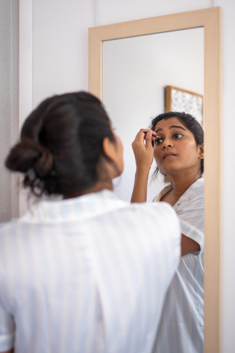 A bride puts eye makeup on in a mirror.