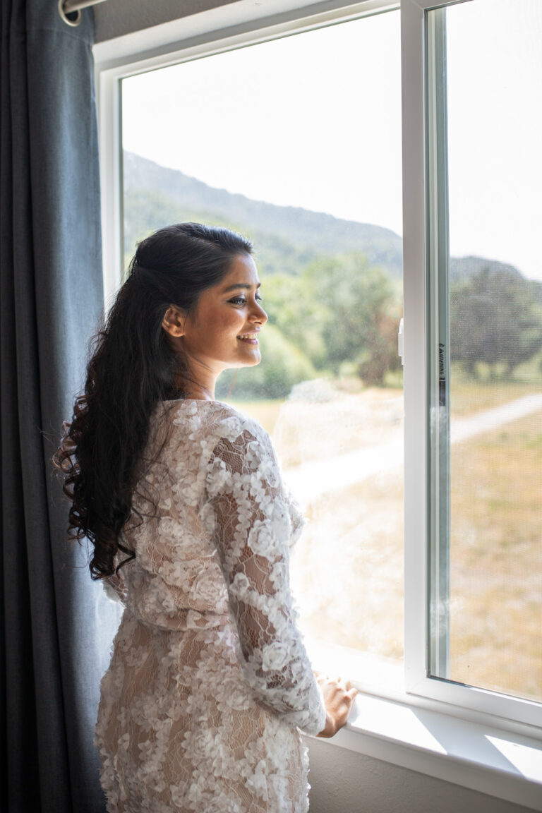 A bride stands in her wedding dress smiling in front of a big window in a house in Big Sur.