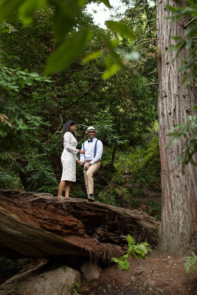 A bride and groom stand together on a log in a forest.