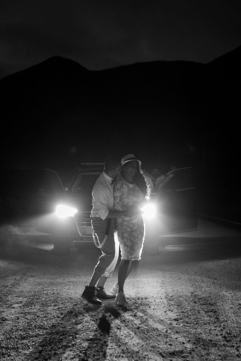 A bride and groom dance in the dark in front of their car with the headlights on.