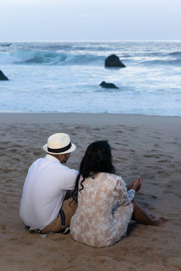 A bride and groom sit in the sand looking out over the ocean after their cliffside wedding in Big Sur, California.