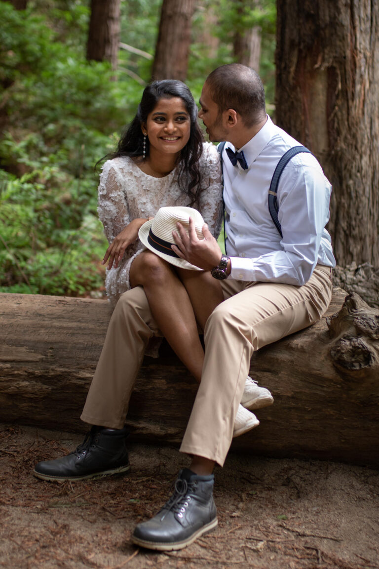 A bride and groom sit on a downed tree log while the groom whispers in the bride's ear and she smiles.