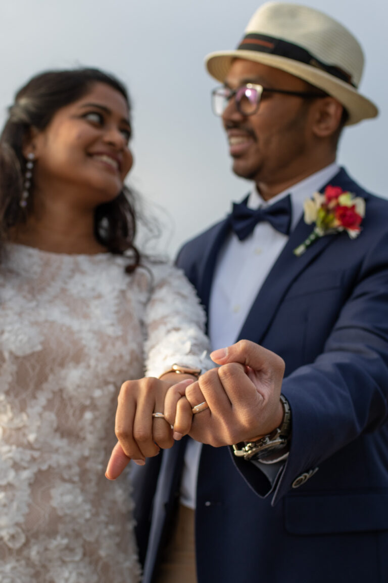 A bride and groom hold out their hands showing off their new wedding bands while smiling at each other.