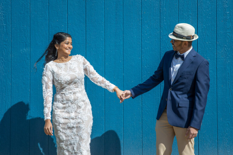 A bride and groom stand in front of a blue building holding hands and smiling at each other.