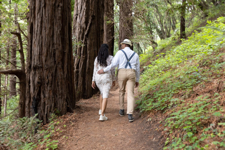 A groom walks with his hand on his bride's lower back as they stroll through the forest together.