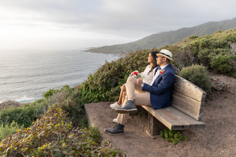 A bride and groom sit on a wooden bench overlooking the ocean after their cliffside wedding in Big Sur.
