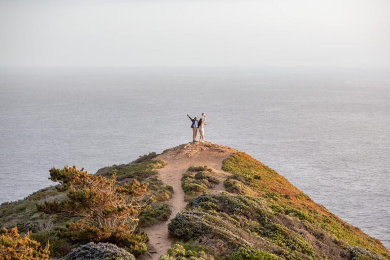 A bride and groom stand on top of a cliff with their hands in the air celebrating after their wedding in Big Sur.