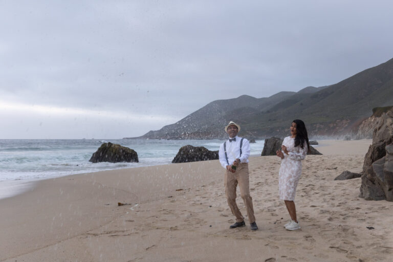A groom sprays champagne while his bride stands next to him on a beach in Big Sur.