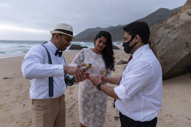 A groom pours champagne for his bride and his best friend, the officiant while standing on the beach in Big Sur.