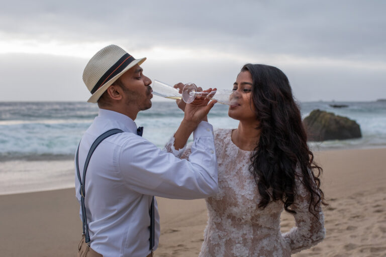 A bride and groom interlock their arms as they sip champagne from glasses on the beach in Big Sur.