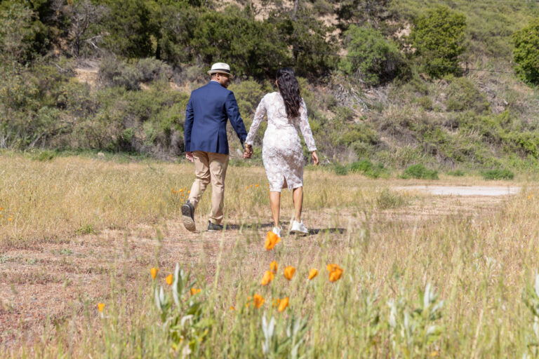 A bride and groom walk hand in hand along a dirt path with orange poppies in the foreground.