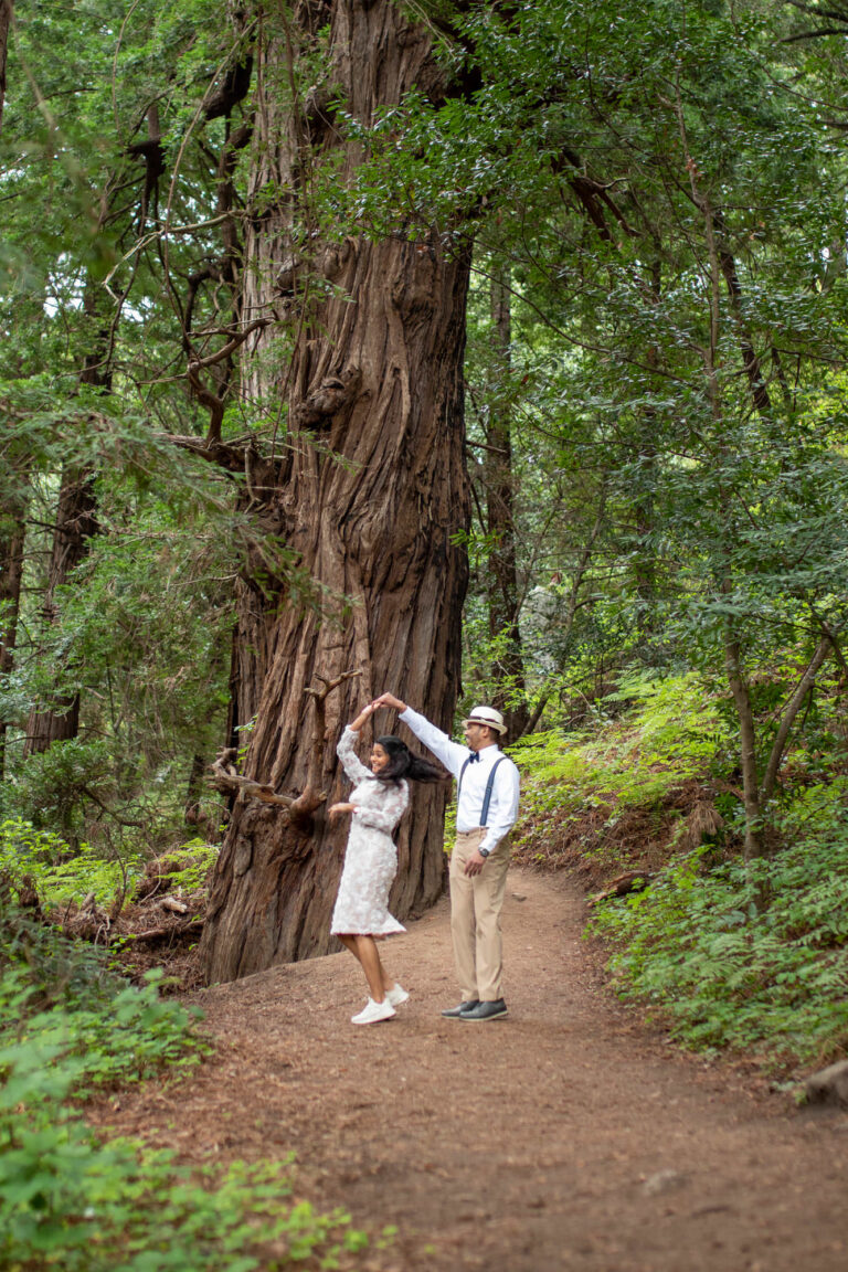 A groom spins his bride as they stand on a dirt path in a forest in Big Sur.
