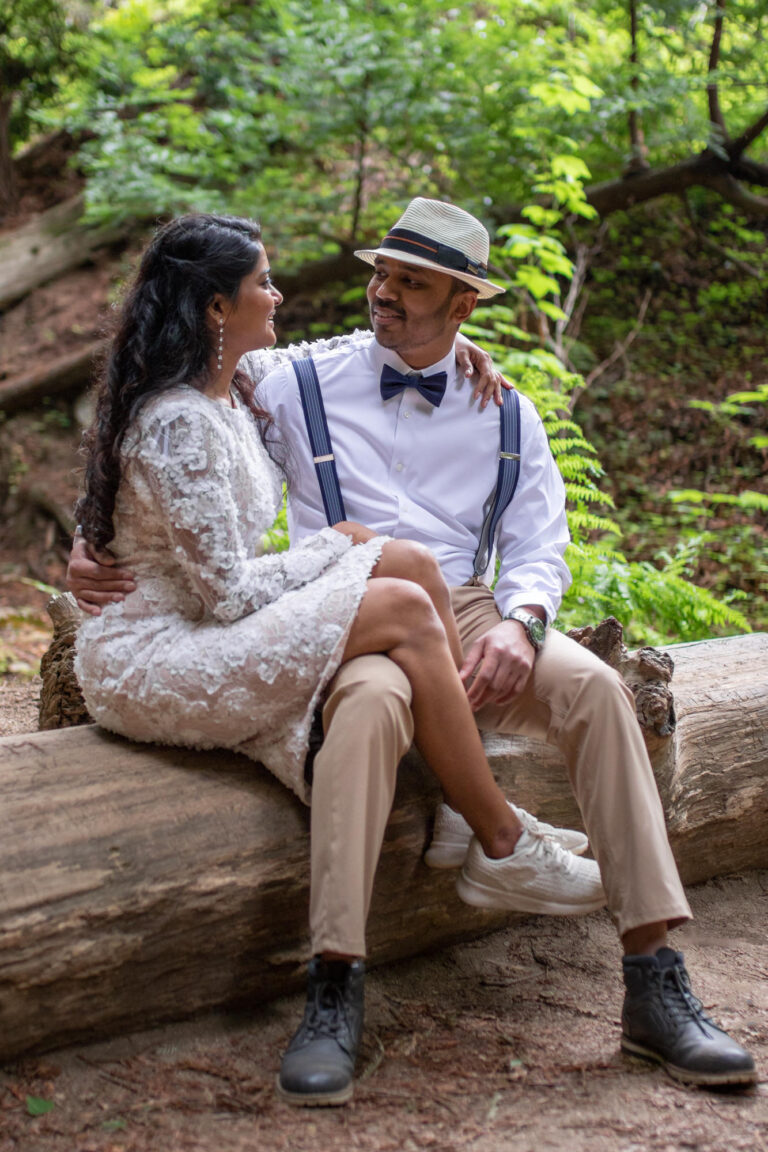 A bride sits on a log with her groom and her legs over his leg as they smile at each other.