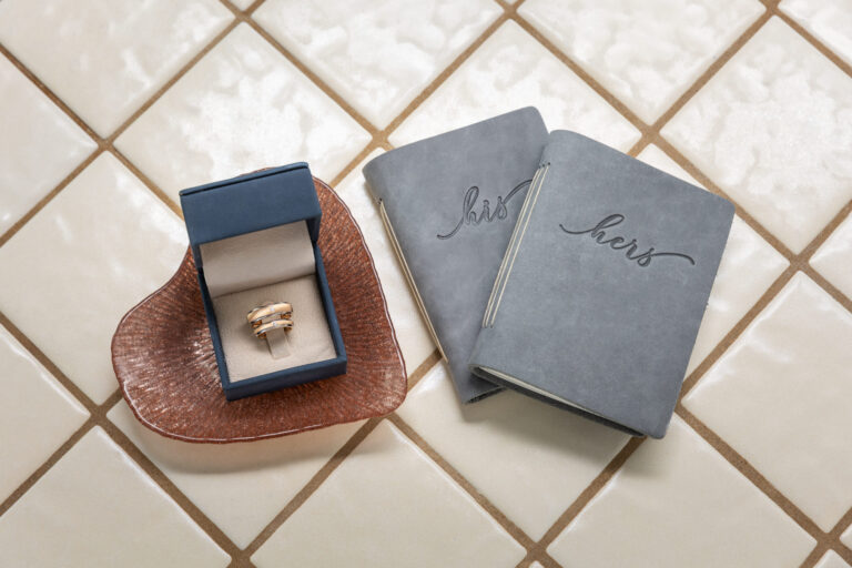Two gray vow books that say "his" and "hers" sit on a tiled floor with a ring box with wedding rings next to them.
