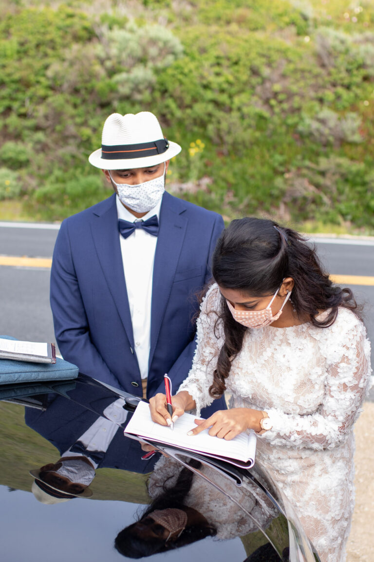 A bride and groom stand next to a black car, signing their marriage license on their wedding day.