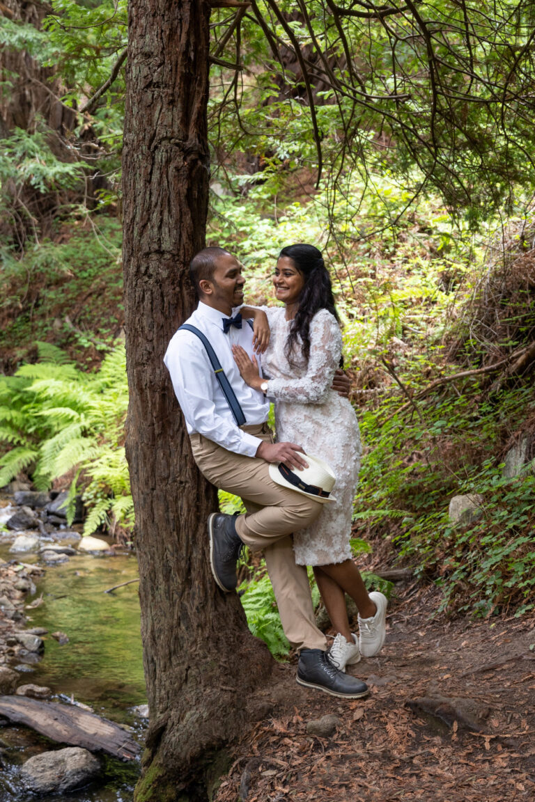 A groom leans against a tree while his bride leans against him, smiling.