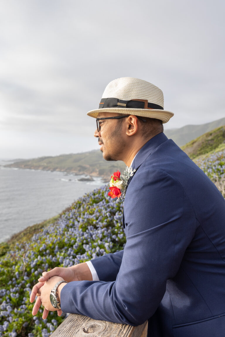 A groom leans over a wooden railing, looking out over the ocean on his cliffside wedding day.