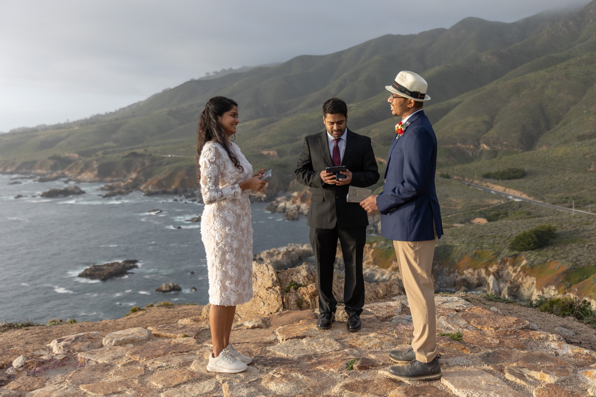 A bride and groom smile at each other while the officiant reads during their cliffside wedding ceremony.