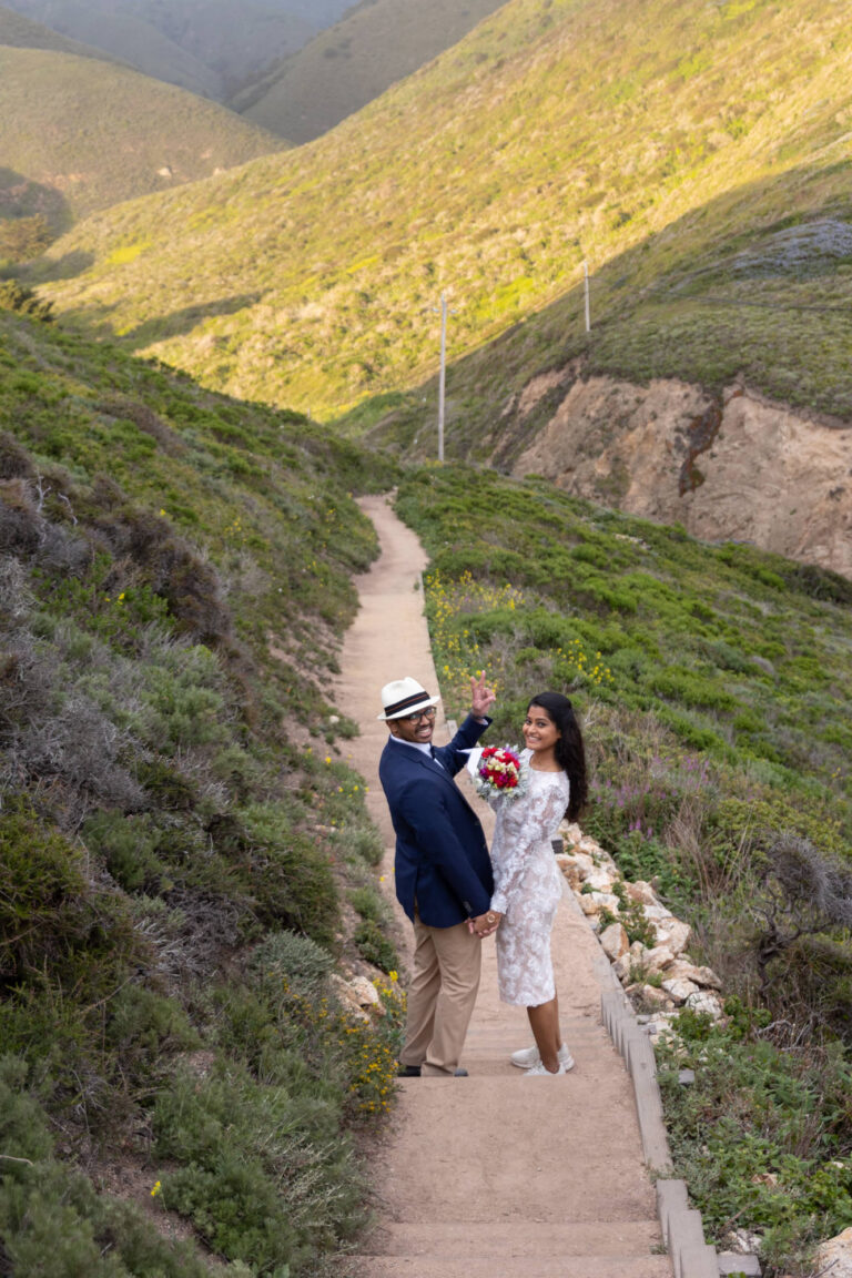 A bride and groom smile at the camera as they walk down stairs in Big Sur.