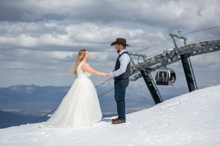 A bride and groom stand facing each other holding hands as the gondola lift comes up behind them.