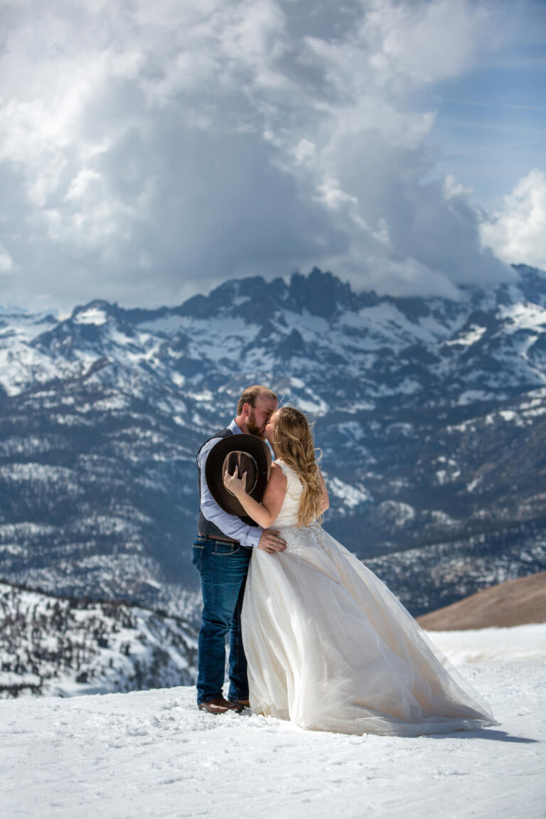 A bride and groom kiss while standing in the snow with mountains behind them and the bride holds the grooms hat in her hand.