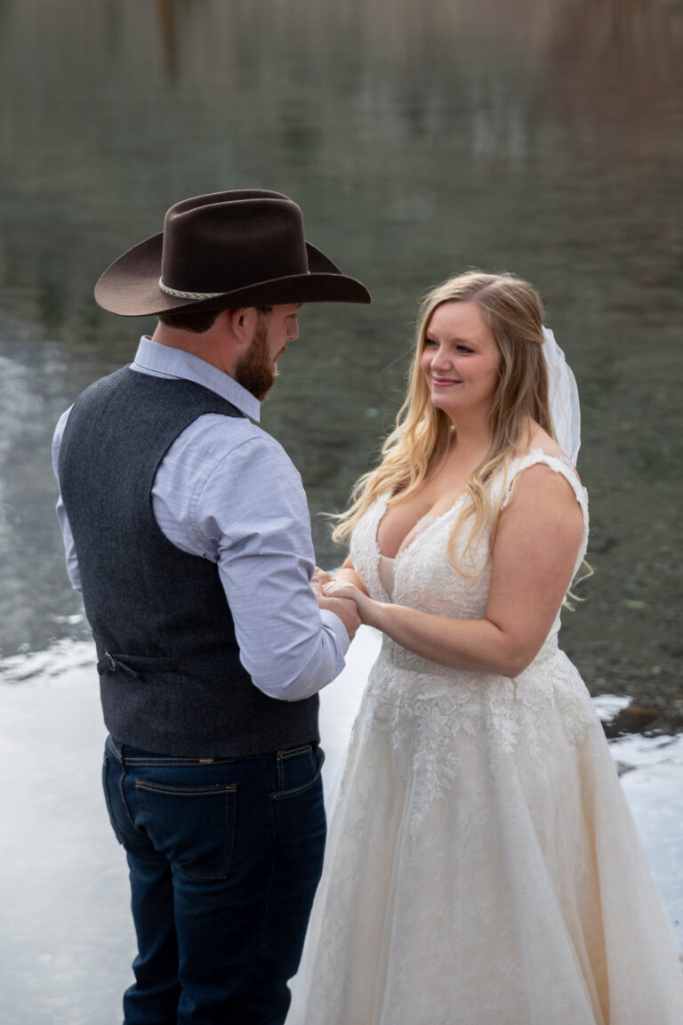 A bride and groom stand holding hands and smiling next to Convict Lake on their wedding day.