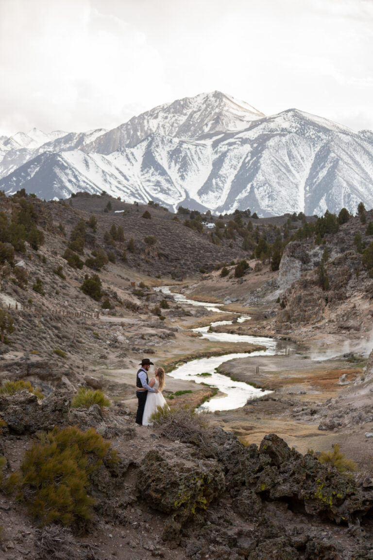 A bride and groom stand with snowy mountains and a river behind them.