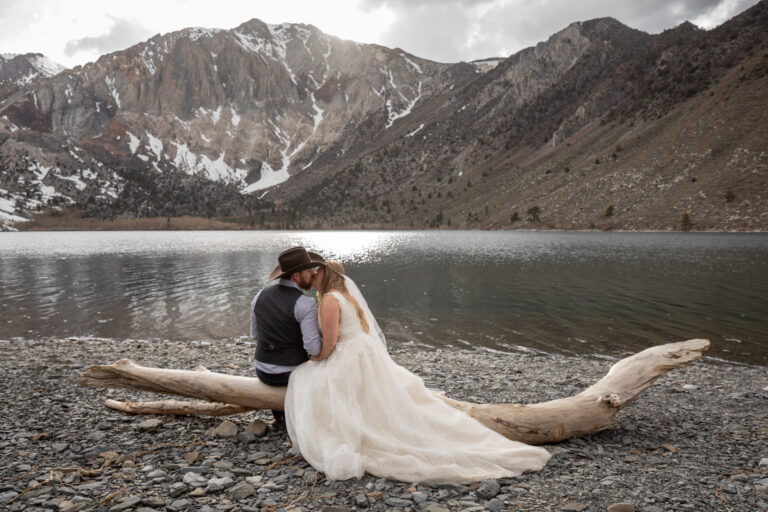 A bride and groom sit on a log at Convict lake beach kissing.