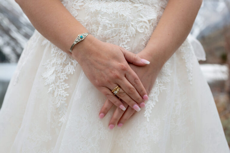 A bride stands with her hands in front of her with the focus on her wedding ring and a bracelet on her wrist.