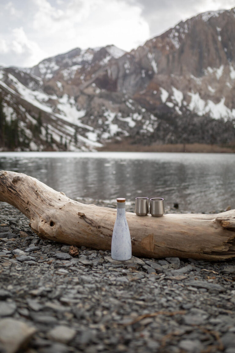 A white bottle is on the beach with two stainless steel cups on a log behind it.