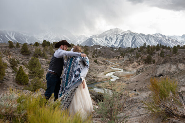 A groom wraps a blue blanket around his bride with the mountains behind them.