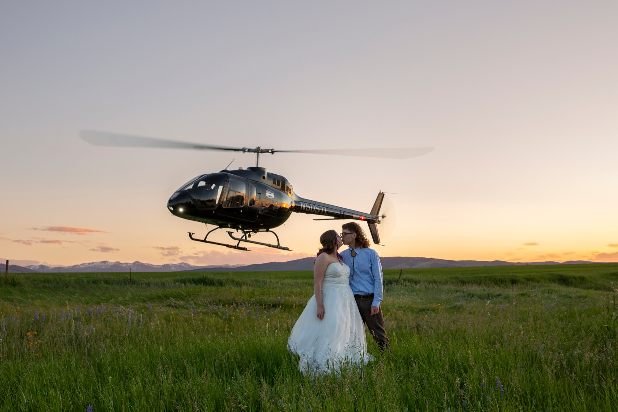 Where to Elope: How to Choose the Perfect Elopement Location | Scenic Vows couple standing in wildflower meadow beside helicopter during adventure elopement