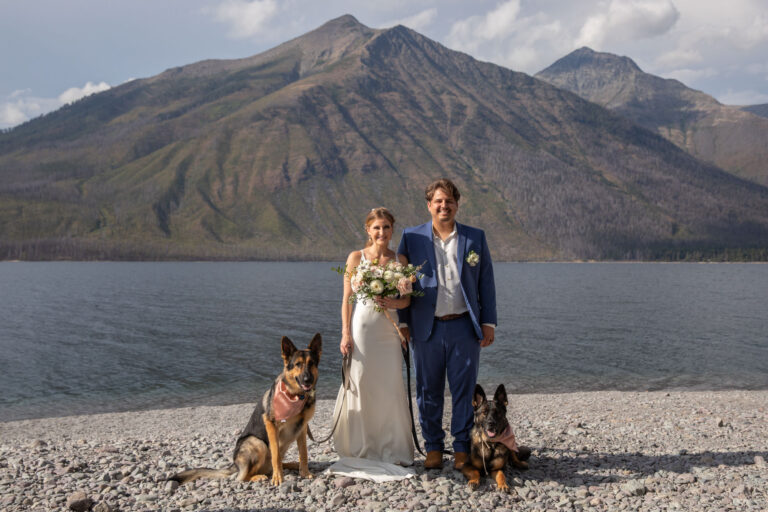 A bride and groom stand next to each other, smiling at the camera with their two dogs on either side of them.