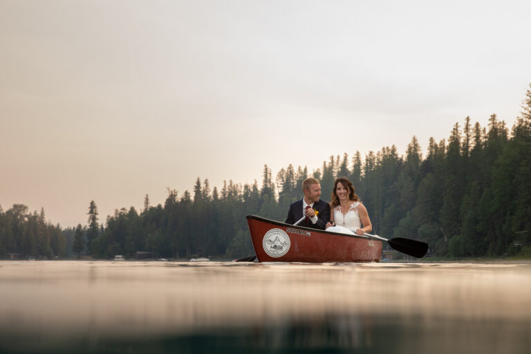 A bride and groom sit in a canoe side by side paddling on Lake McDonald after they elope in Montana.