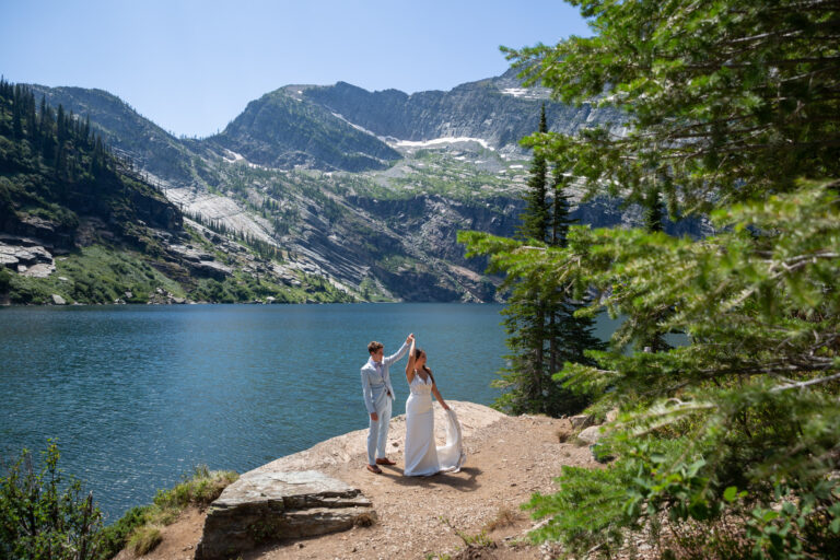A groom twirls his bride under his arm as they stand next to a lake in Montana with green trees all around them.