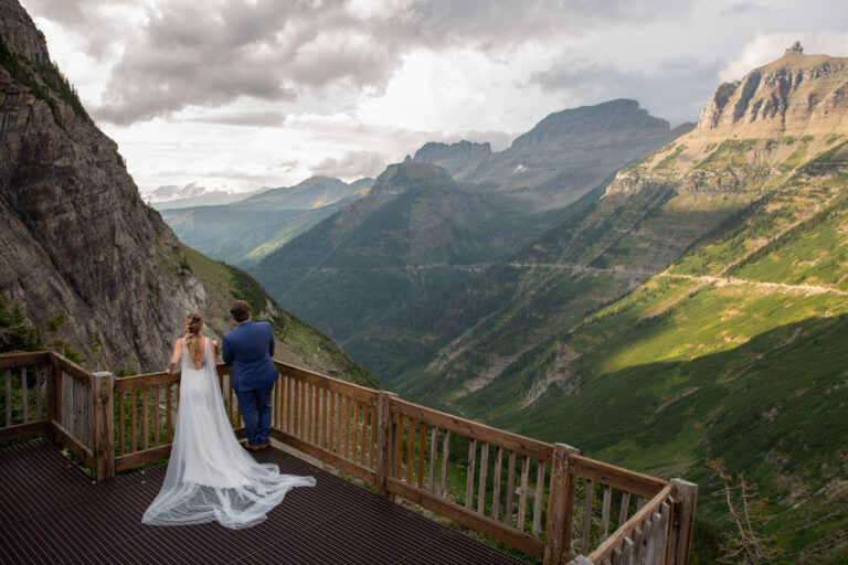 A bride and groom lean against a wooden fence as they stand on a metal platform looking out over the mountain views in Montana.
