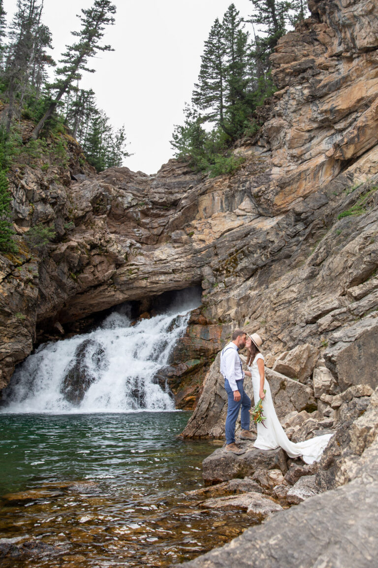 A bride and groom stand next to a waterfall in Montana on a rock kissing.