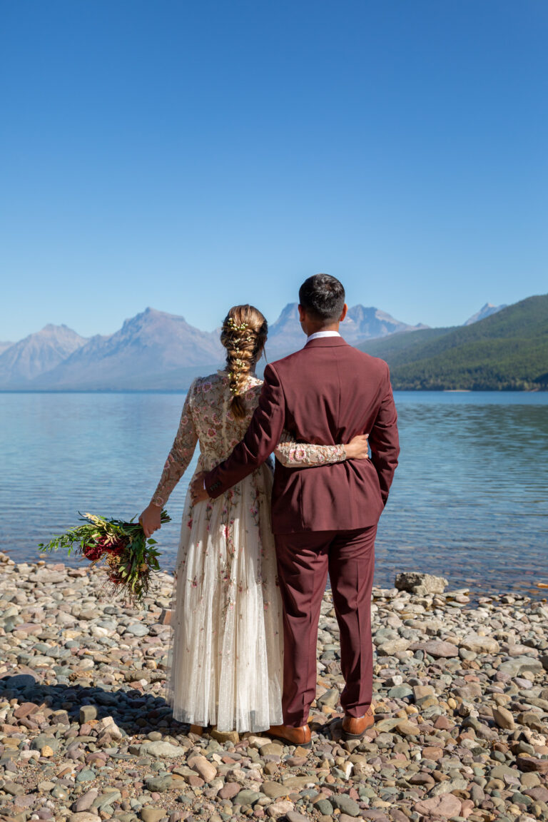 A bride and groom stand with their arms around each others waist while they look out over Lake McDonald in Montana.