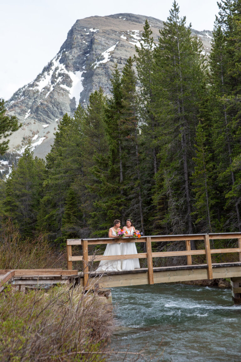 Two brides lean against a wooden railing on a wooden bridge with trees and a snowy mountain behind them