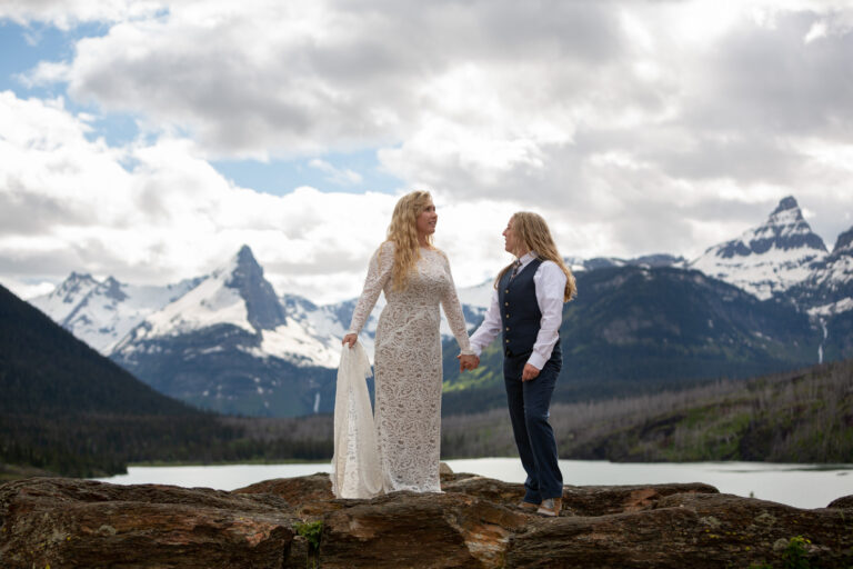 Two brides stand holding hands looking around at the mountain scenery around them.