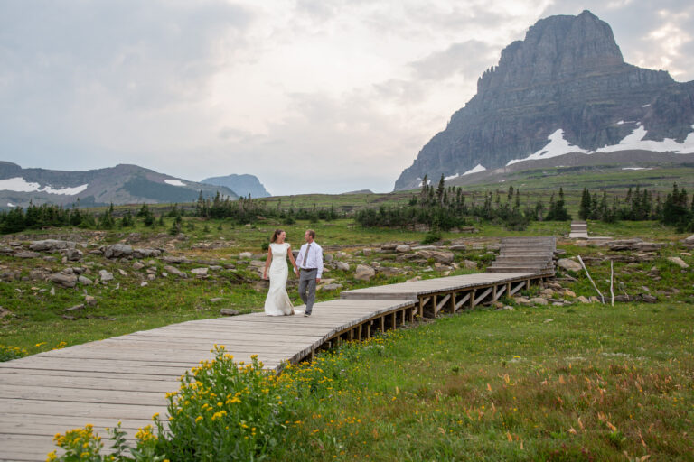 A bride and groom walk hand in hand on a wooden boardwalk in a meadow of yellow flowers with mountains in the background.