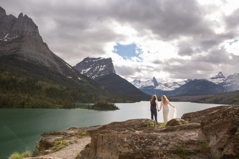 Two brides stand holding hands looking over a lake in one of our favorite places to elope in Montana, Glacier national Park.