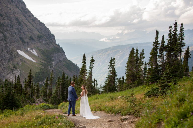 A bride and groom holding hands and standing on a dirt trail looking back over their shoulders smiling at the camera.